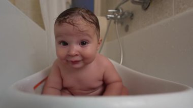 Küvette bebek, tuhaf bir ifade, ıslak saçlar, banyo zamanı, duyusal oyundan zevk almak, dik oturmak, evdeki hijyen rutini sırasında çevreyi keşfetmek.