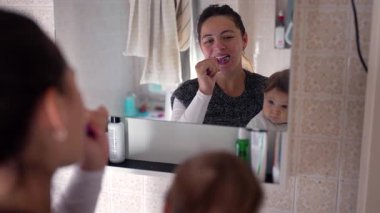 Anne banyoda dişlerini fırçalarken bebeğe bakıyor, kişisel bakımla özenli ebeveynliği birleştiriyor, günlük rutinde sıcak ve şefkatli bir aile etkileşimi kuruyor.