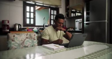 Afrika kökenli bir adam mutfak masasında oturuyor, stresli ve bunalmış, elleri alnında, laptopta çalışıyor, işiyle boğuşuyor, finansal problemler yaşıyor, uzaktan iş stresi yaşıyor.
