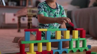 Renkli bir blok inşa etmeye odaklanan çocuk, yaratıcı oyun ve problem çözme ile meşgul, kapalı bir evde odaklanmış bir anın tadını çıkarıyor.