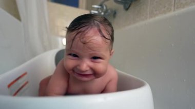 Bebek küvette, neşeli bir gülümsemeyle, başını çevirerek, banyo zamanı eğlencesi, ıslak saçlar, neşeli ve rahat sıcak ev ortamında duyusal hijyen rutini sırasında.