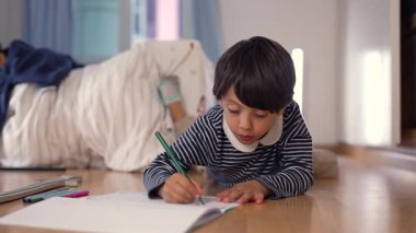 Çocuk yere uzanmış, yeşil bir kalemle çizim yapıyor, ev ortamında odaklanmış ve yaratıcı, çocukluk hayalinin bir anını yakalıyor.