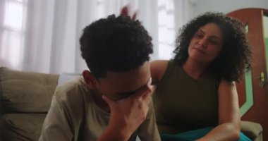 Kanepede oturan genç bir çocuk, duygusal olarak zorlanıyor ve yüzünü eliyle kapatıyor, annesi teselli edici bir jest, aile empatisi ve özenle destek veriyor.