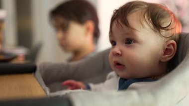 Bebek minderli bir sandalyede oturuyor, gözleri sonuna kadar açık, meraklı bir şekilde çevreyi keşfediyor, arka planda büyük kardeş, evde sessiz bir gözlemle meşgul.