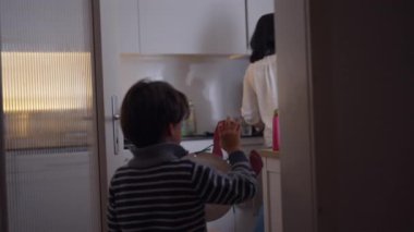 Genç bir çocuk mutfakta el sanatları projesine giriyor, anne mutfak tezgahının yanında bebeği tutuyor, günlük aile ilişkileri ev işleri sırasında.