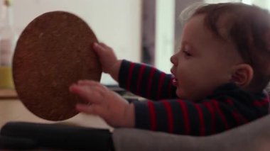 Bebek yüksek sandalyede oturuyor, bir testere tutuyor ve garip bir ifadeyle yalıyor, duyusal etkileşim yoluyla dokuyu keşfediyor, erken gelişim evresi, kapalı bir aile ortamı.
