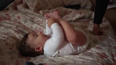 Bebek çiçekli yatakta uzanıyor, iki eliyle ayaklarını tutuyor, neşeli ve meraklı, yumuşak ve rahat bir yuva kuruyor.