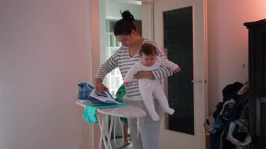 Anne kıyafetleri ütülerken ve bebeği tutarken birden fazla iş yapar, ev işlerini ebeveynlikle dengeler, otantik aile anı, bakım, günlük ev hayatı, samimi ifade