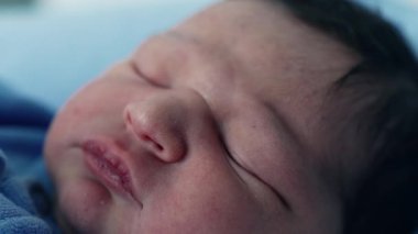 Yeni doğmuş bir bebeğin huzur içinde uyuduğu, narin yüz hatlarının altını çizen, yumuşak cilt ve huzurlu yüz ifadesi, masumiyeti, sükuneti ve yeni bir hayatı temsil eden mavi bir battaniyeye sarılı.