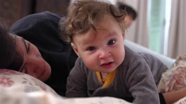 Bebek kameraya doğrudan meraklı bir ifadeyle bakarken anne yan yana uzanmış, meşgul ve dikkatli, samimi aile anı, kapalı alanda yakın bağ, erken çocuk gelişimi