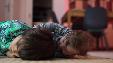 Bebek ve büyük kardeş yerde yatıyor, neşeli bir etkileşim içinde, bebek şakacı bir şekilde gülümserken kardeş saçı çekiyor, bağ kuruyor, aile bağlarını sıcak bir ortamda besliyor. 