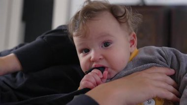 Bebek annesinin kolunda yatarken siyah kayışı çiğniyor, odaklanmış ve meraklı ifade, ağzıyla keşfetmek, kapalı alanda eğlenceli bir aile anı, dokunsal bir keşif