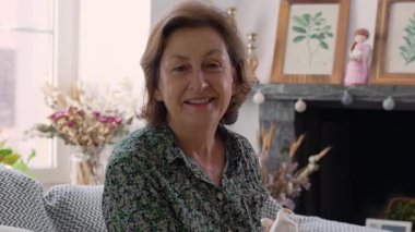 Gülümseyen yaşlı kadın çiçeklerle süslü bir oturma odasında oturuyor, sıcaklığını ve mutluluğunu iletiyor.
