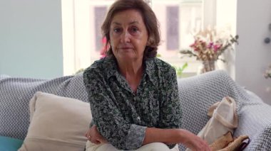 Rahat bir kanepede, rahat, aydınlık bir oturma odasında oturan yaşlı bir kadın. Yastıklarla ve ev dekorlarıyla çevrili sakin bir ifadesi var.