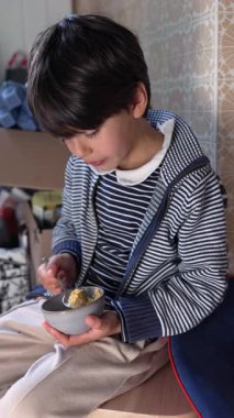 Küçük kaseden yemek yiyen küçük bir çocuk, odaklanmış bir ifade, sıradan bir iç mekan anı, basit bir günlük rutin, bağımsız yeme alışkanlığı, çocukluk alışkanlıkları, rahat yemek saatinin samimi bir şekilde yakalanması.