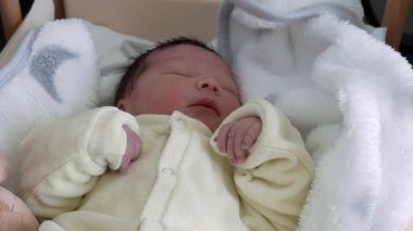 Yeni doğan bebek, yumuşak, beyaz bir battaniyeye sarılı, tahta bir beşikte huzur ve masumiyet dolu bir anı yakalıyor.