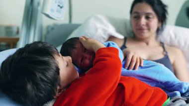 Mavi tulumlu yeni doğmuş bebek hastane yatağında ablasıyla sarmaş dolaş, anne de arka planda sevgiyle izliyor. Şefkatli bir aile anın altını çiziyorum.