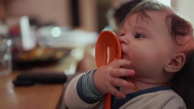 Bebek portakallı kaşık tutuyor, yanağa bastırıyor, gevşemiş ifade, yemek saatinde sandalyeye oturuyor, duyusal keşiflere ince odaklanıyor, neşeli bir evcil an.
