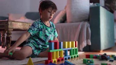 Genç bir çocuk yerde renkli bir blok inşa ediyor, yaratıcı oyun ve problem çözme üzerine odaklanmış, sıcak bir ev ortamında iç mekan aktivitesinin keyfini çıkarıyor.