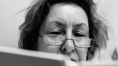 Tek renkli, yaşlı bir kadının gözlük taktığı, okumaya odaklanmış bir fotoğrafı. Yüzündeki bilgelik çizgileri yılların deneyimini yansıtıyor.