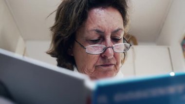Gözlük takan yaşlı kadın derinden kitap okumaya odaklanmış. Yoğun konsantrasyon. Yazılı materyali özümsemeye olan bağlılığını vurguluyor.