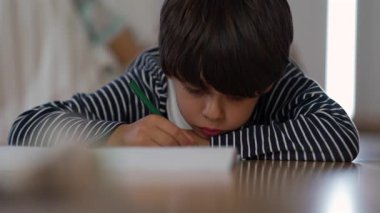 Yerde yatan genç bir çocuk, yeşil kalemle çizerken, konsantrasyonunu, yaratıcılığını ve sanatsal veya öğrenme aktivitelerine olan bağlılığını ifade ediyor.