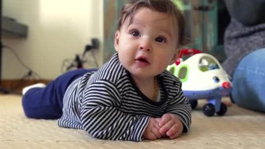 Bebek yerde neşeli bir ifadeyle yatıyor, kolları desteklenirken geniş bir şekilde gülümsüyor, mutluluk ve erken gelişim nişanı sergiliyor, arka planda görünen oyuncak uçak