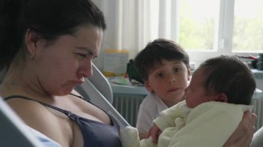 Anne, yeni doğmuş bebeğine şakacı bir şekilde surat asarken, büyük kardeş merakla bakıyor. Komik etkileşim aile içindeki neşe ve sevgiyi vurgular, neşeli ve sevgi dolu bir aile anını yakalar.