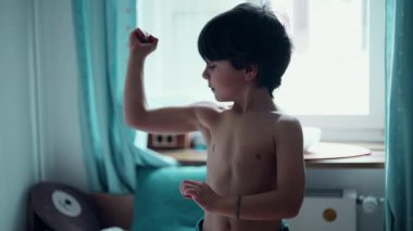 Şirin, üstsüz, kollarını açan genç bir çocuk, yatak odasında gücünü gösteren 5 yaşında, neşeli, beyaz bir çocuk. Çocuk kaslarını gösteriyor.