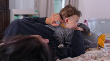 Anne bebeğin kulağına hafifçe dokunurken bebek dik, odaklanmış ve meraklı bir şekilde oturur hassas bir bağ, aile bağı ve erken çocukluk etkileşimi gösterir.
