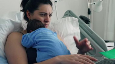 Anne, dizüstü bilgisayarında çalışırken yeni doğmuş bebek tulumuyla hastane yatağında birden fazla iş yapıyor. Erken ebeveynliğin dengesini ve sorumlulukların sorumluluğunu yakalıyor.