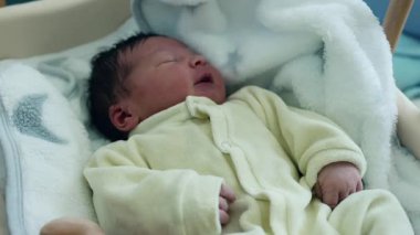 Yeni doğmuş bebek, yumuşak, sarı bir tulum giymiş, tahta bir beşikte huzur içinde uyuyor ve tüylü beyaz bir battaniyeye sarılıp huzurlu bir dinlenme ve masumiyet anı yakalıyor.