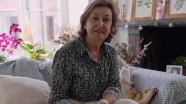 Güzel dekore edilmiş bir oturma odasında kanepede oturan yaşlı kadın, düşünceli bir ifadeyle kameraya bakıyor, bitkiler ve çerçevelenmiş sanat eserleriyle çevrili.