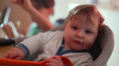 Bebek yüksek sandalyede oturuyor, elinde büyük bir turuncu plastik kaşık tutuyor, ilginç bir şekilde çatal bıçakları keşfediyor ve dokunuyor, yemek saatinde duyusal bir oyun oynuyor.