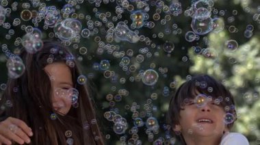 Çocuklar 1000 fps 'te yavaş çekimde baloncukların keyfini çıkarıyorlar, sayısız renkli kabarcıklarla çevrili, dışarıda eğleniyor ve gülüyorlar.