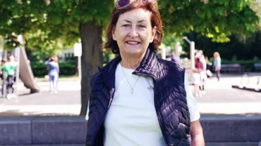 Açık hava parkında gülümseyen yaşlı bir kadın, günlük beyaz gömlek ve siyah yelek giyiyor, arka planda ağaçlar ve oyun ekipmanları var, güneşli bir gün.