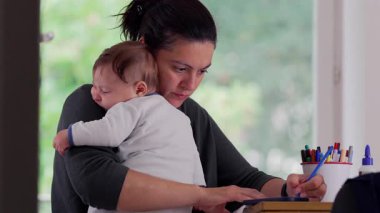 Anne bebeğini masada yazarken kucağında tutuyor, sakin ve besleyici bir ofis ortamında işini tamamlamaya odaklanıyor, bakımı verimlilikle dengeliyor.