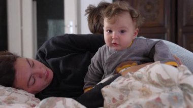 Bebek yatağında dinlenirken annesinin koluna oturuyor, neşeli bir ifadeyle gülümsüyor, sıcak bir aile anı, sevgi dolu bir ev içi bağ kurma zamanı, huzurlu ve sıcak bir ev ortamı.