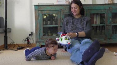 Bebek yerde sürünüyor, erken gelişim hareketleri gösteriyor, anne oyuncak uçakla yakınlarda oturuyor, meşgul ve gülümsüyor, ev ortamında gündelik bağ kuruyor.