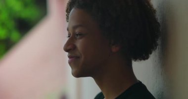 Olumlu bir ifadeyle gülümseyen, duvara dayanan, Afrika kökenli, açık hava manzaralı, mutluluk ve memnuniyet dolu, yakın çekim profilli bir çocuk.