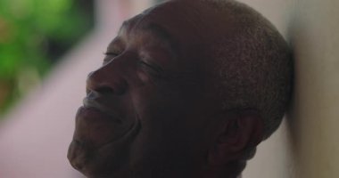 Afrika kökenli yaşlı bir adam, sıcak bir gülümseme, başını yaslanmış, bir duvara yaslanmış, açık havada canlı yeşillik, coşkulu bir sevinç, sıcaklık, ve bir karşılama davranışı ile