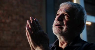 Elleri sımsıkı bağlı dua eden yaşlı bir adam, bir inanç ve yansıma anında yukarıya bakıyor, bir haçın yanında ruhani bir ortamda umut ve sadakati iletiyor.
