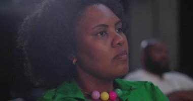 Seyirciler arasında renkli bir kolye takan bir kadın, dini bir etkinlik sırasında ciddi bir ifadeyle dikkatle dinliyor.