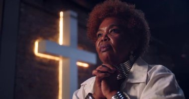 Yaşlı bir kadının dua ederken ellerini sıkarak gözlerini açması umut dolu ve inançlı bir ifadeyle, ruhani bir ortamda ışıldayan bir haçla aydınlanmış.