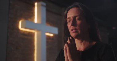Kilisede dua eden orta yaşlı bir kadın, gözleri kapalı, elleri sımsıkı bağlı, arka planda parlayan haç, derin manevi yansıma, inancı, umudu ve bağlılığı simgeliyor.
