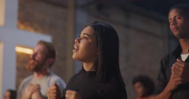 El sıkışık ibadet eden genç kadın, derin ruhani bağlılık, Hristiyan inancı, dua eden an, toplum birliği, arka planda aydınlanmış haç