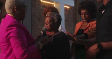 Bir dua grubunda Afrika kökenli Amerikalı bir kadın. Elleri destek grubunda. Etrafı duygusal, ruhsal bir ortamda çeşitli insanlarla çevrili.