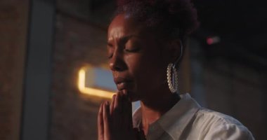 Elleri sımsıkı bağlı dua eden genç siyahi bir kadının yan profili, gözleri derin bir yansımayla kapalı, tuğla arka planda yumuşak bir odak, inanç ve maneviyatı simgeliyor.