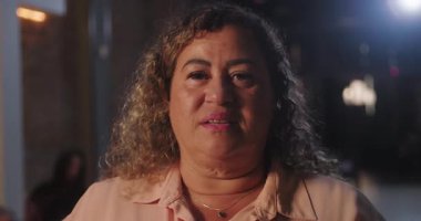 Stüdyo ortamında duran kıvırcık saçlı Latin kadın, kamerayla doğrudan göz teması kuruyor, sakinliğini ve duruşunu yansıtan anlamlı bir yüz ifadesi sergiliyor.