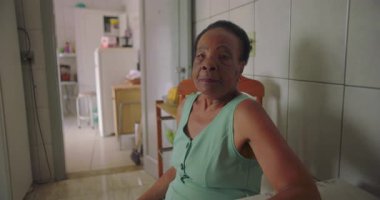 Rahat bir ev mutfağında yemek masasında oturan yaşlı Afro-Amerikalı kadın, hafif bir kızgınlıkla iç çekiyor, yalnızlığı ve duygusal derinliği yansıtıyor.